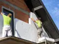 Pracownicy montują styropian na ścianie budynku, realizując termomodernizację.