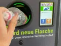 Ręka wrzuca butelkę do automatu, który jest częścią systemu kaucyjnego w Niemczech. Na ekranie widać flagi, symbol butelki i opcje zwrotu kaucji.