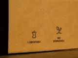 Na brązowym opakowaniu widnieje znak opakowanie biodegradowalne. Obok symbolu jabłka i napisu "COMPOSTABLE" znajduje się symbol kiełkującej rośliny z napisem "BIO DEGRADABLE".