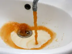 Brązowa woda z kranu, prawdopodobnie z powodu manganu w wodzie pitnej, tworzy rdzawy osad w białej umywalce.