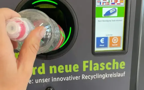 Ręka wrzuca butelkę do automatu, który jest częścią systemu kaucyjnego w Niemczech. Na ekranie widać flagi, symbol butelki i opcje zwrotu kaucji.