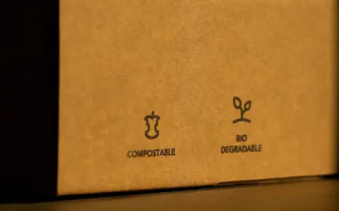 Na brązowym opakowaniu widnieje znak opakowanie biodegradowalne. Obok symbolu jabłka i napisu "COMPOSTABLE" znajduje się symbol kiełkującej rośliny z napisem "BIO DEGRADABLE".