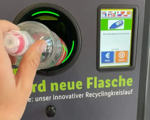 Ręka wrzuca butelkę do automatu, który jest częścią systemu kaucyjnego w Niemczech. Na ekranie widać flagi, symbol butelki i opcje zwrotu kaucji.