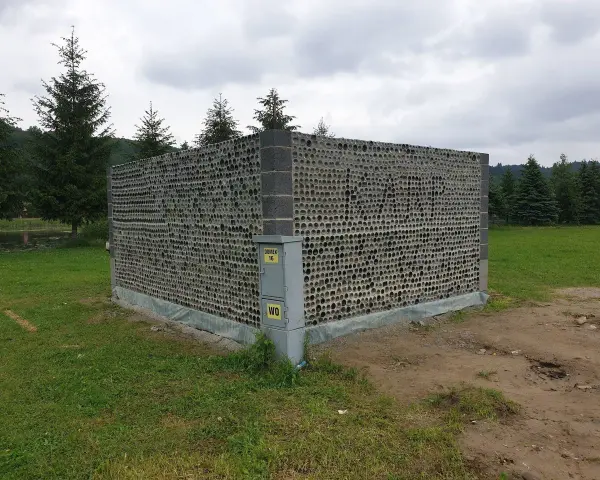 Nietypowy domek z butelek plastikowych, zbudowany z tysięcy szklanych opakowań, otoczony zielenią i drzewami.