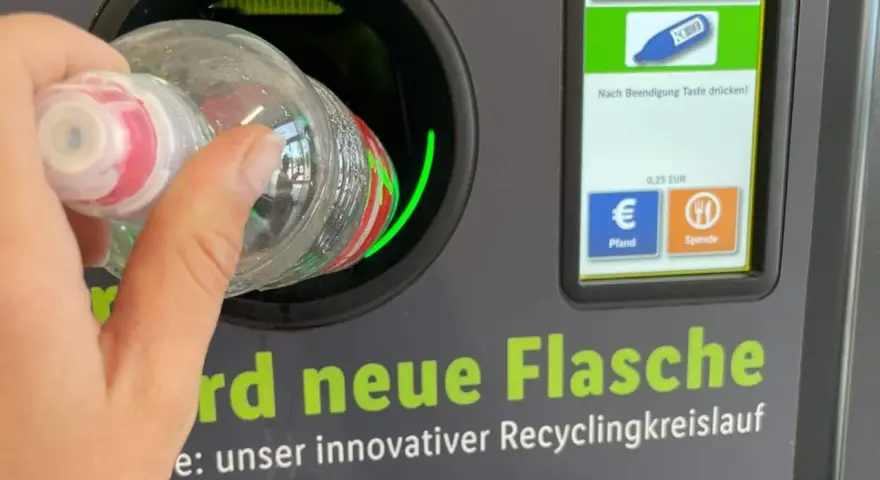 Ręka wrzuca butelkę do automatu, który jest częścią systemu kaucyjnego w Niemczech. Na ekranie widać flagi, symbol butelki i opcje zwrotu kaucji.
