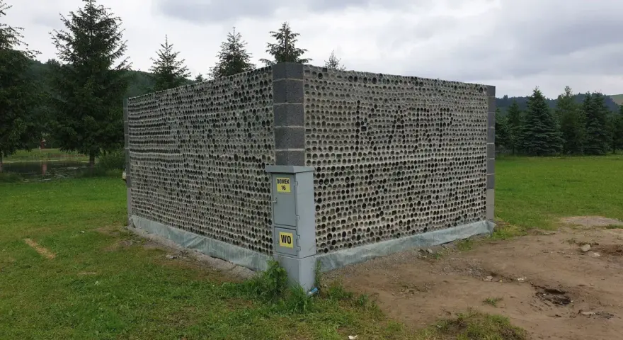 Nietypowy domek z butelek plastikowych, zbudowany z tysięcy szklanych opakowań, otoczony zielenią i drzewami.