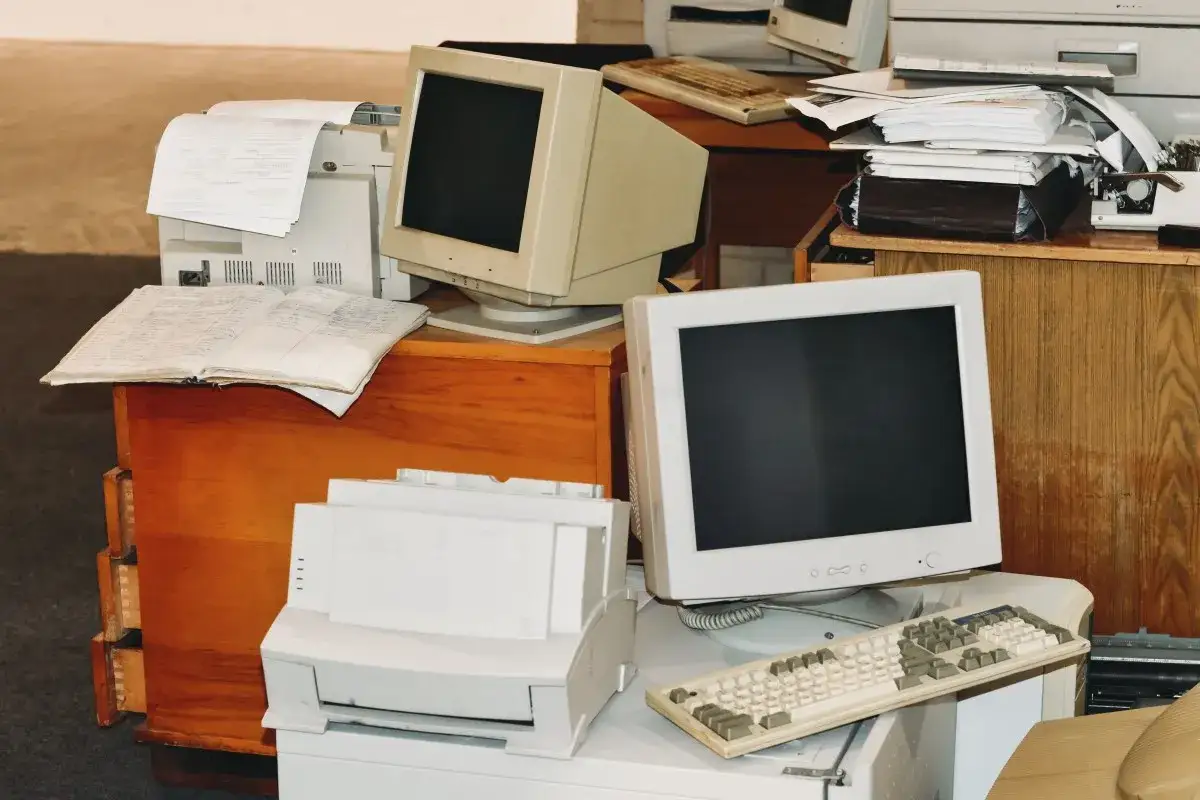 Stary komputer stacjonarny, drukarka i otwarty zeszyt leżą na drewnianej szafce. Obok stoi kolejny monitor i klawiatura.